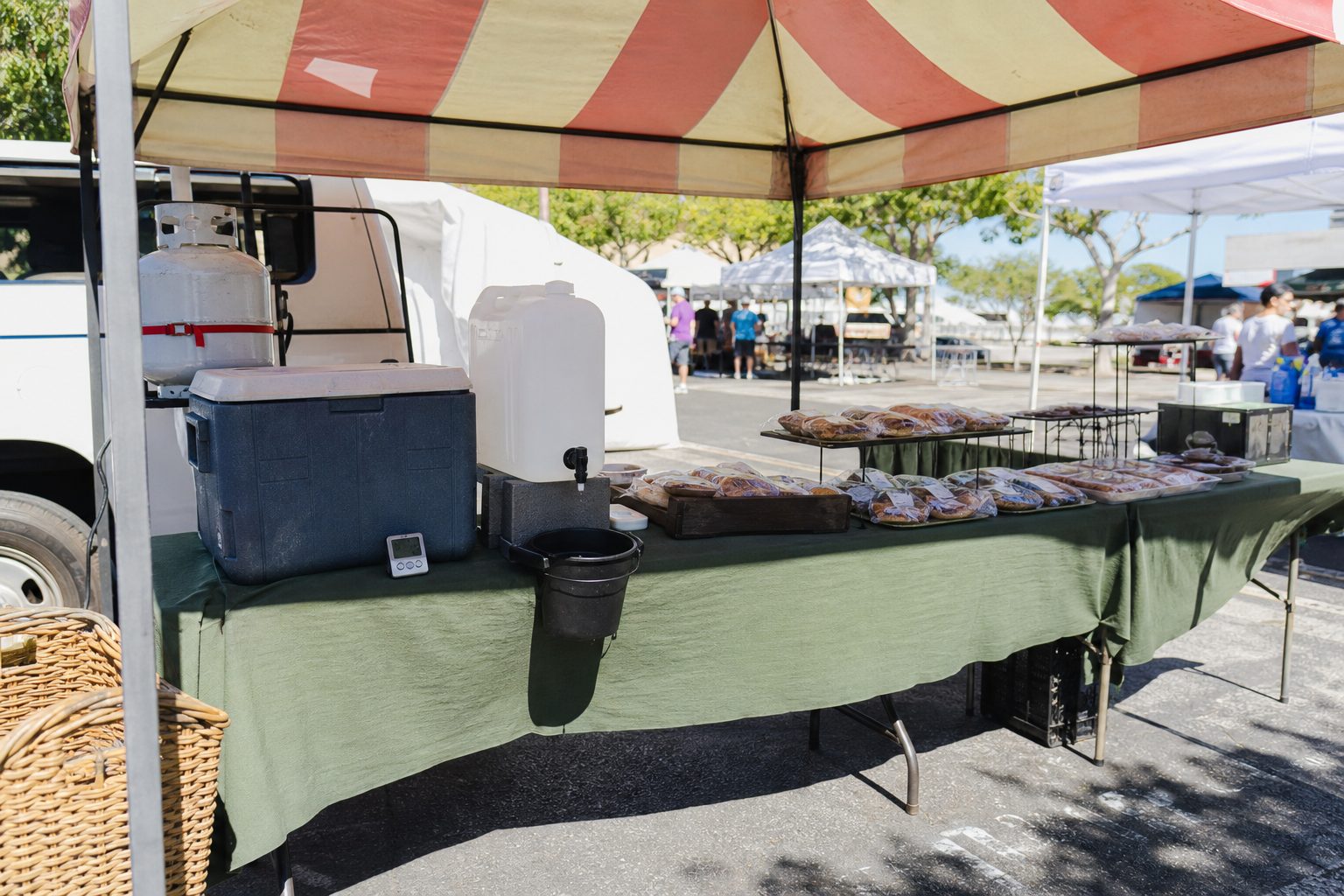 Prepared food stall with a cooler, thermometer and handwashing jug under a striped canopy in midday light